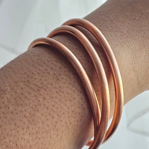 May include: Four copper bangles stacked on a person's wrist. The bangles are smooth and shiny.