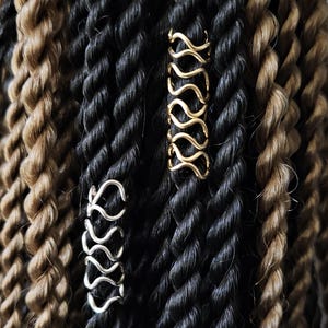 May include: Two hair cuffs, one silver and one gold, are used to hold back braids. The braids are a mix of black and brown.