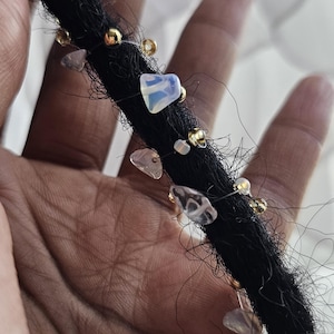May include: A black dreadlock hair extension adorned with a spiral of small gold-toned metallic beads and various translucent, irregular-shaped gemstone chips in shades of pale pink, clear, and iridescent white, secured with a thin clear filament.