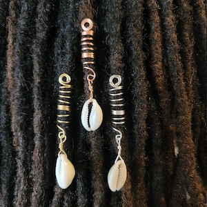 May include: Three gold, silver, and copper dreadlock beads with white cowrie shells hanging from them. The beads are coiled and have a decorative design.