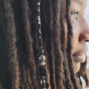 May include: A beaded dreadlock accessory featuring a strand of black, silver, and clear beads. The beads include various shapes and sizes, with some having a textured or patterned appearance. The accessory is shown against a backdrop of dark brown dreadlocks.