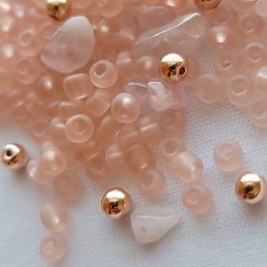 May include: A collection of small, translucent pink beads and gold beads. The beads are scattered on a white surface.