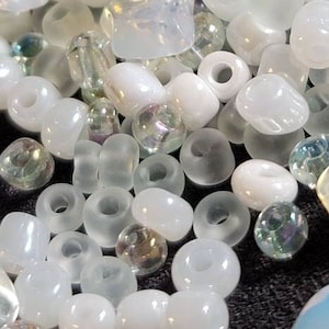 May include: A close-up shot of a collection of white and iridescent beads. The beads vary in shape and size, including round, oval, and cube-shaped. Some beads have a pearlescent sheen, while others are clear, creating a textured and visually appealing display.