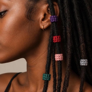 May include: A side profile view of a person with dark locs adorned with colorful crochet hair beads. The beads are in shades of purple, red, pink, gray, and green. The person is wearing a black tank top.