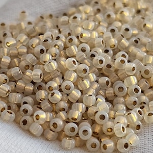 May include: A pile of small, cylindrical beads with a translucent, off-white appearance and golden-colored centers. The beads are scattered on a light gray fabric, creating a textured surface. These beads are ideal for jewelry making and crafts.