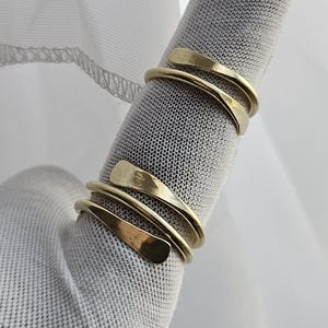 May include: Two gold-colored, spiral-design rings are displayed on a finger. The rings have a smooth, polished finish and wrap around the finger in a layered effect. The background is a soft, white fabric.