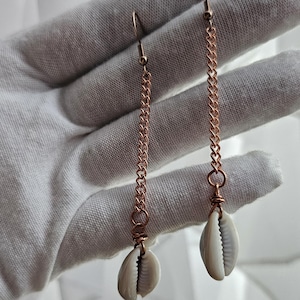 May include: A pair of long drop earrings featuring rose gold-toned metal chains suspended from ear wires. Each chain terminates in a single, off-white cowrie shell secured with matching rose gold-toned wire wrapping at the base of the chain.