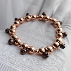 Könnte beinhalten: Ein roségoldenes Armband mit kleinen, runden Glöckchen, die daran hängen. Das Armband besteht aus Metallperlen und liegt auf einer weißen Oberfläche.