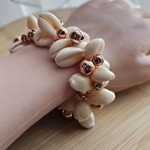 May include: A seashell bracelet with cream-colored shells and copper-colored beads. The bracelet is displayed on a wooden surface, showcasing the natural tones of the shells and the metallic accents.