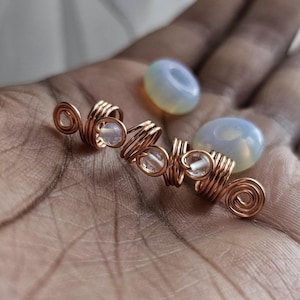 May include: A copper wire hair coil featuring a spiral design with small, clear, round beads integrated into the metal loops, accompanied by two separate, smooth, translucent, pale blue, circular beads with a central hole.