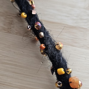 May include: A single black dreadlock extension adorned with a variety of decorative beads in shades of amber, orange, yellow, and metallic gold. The beads are secured along the length of the textured hair strand with thin, dark wire wrapping.