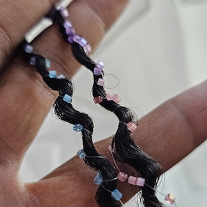 以下が含まれることがあります： 小さな半透明のビーズで飾られた、ウェーブのかかった黒いヘアエクステンションの2本の毛束。1本の毛束には水色と紫のビーズが、もう1本の毛束にはピンクと紫のビーズがあしらわれています。それぞれの毛束の端には、小さな銀色の金属製の輪が付いています。