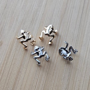 Könnte beinhalten: Vier goldene und silberne Frosch-Anhänger. Die Anhänger sind in Form eines Frosches mit ausgestreckten Beinen. Die Anhänger befinden sich auf einer Holzoberfläche.