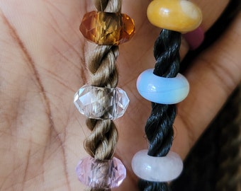 May include: Two braids of hair, one blonde and one black, with colorful beads. The beads are clear, pink, light blue, orange, and dark blue.