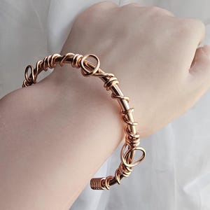 May include: A rose gold wire wrapped bangle bracelet with a unique design. The bracelet is made of multiple strands of wire that are twisted and intertwined to create a textured look.