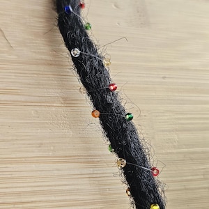 Könnte beinhalten: Eine einzelne schwarze Dreadlock-Verlängerung mit einer spiralförmigen Umwicklung aus dünnem, klarem Faden, der mit kleinen, mehrfarbigen Glasperlen in Rot, Grün, Blau, Gelb und Transparent verziert ist, die gleichmäßig über die Länge verteilt sind.