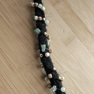 Può includere: Una singola extension per dreadlock nera decorata con una serie di piccole perline nelle tonalità verde chiaro, crema e oro metallizzato. Le perline sono intervallate lungo la lunghezza della ciocca di capelli scura e testurizzata.