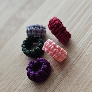 May include: A collection of six handmade crochet rings in various colors. The rings are in shades of gray, green, purple, pink, and burgundy. The rings are arranged on a light wood surface.