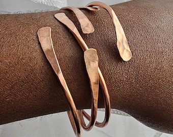 May include: Three hammered copper bracelets stacked on a person's wrist. The bracelets are a warm copper color and have a slightly irregular shape.