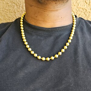 Op de afbeelding: Een gouden ketting met ronde kralen. De ketting wordt gedragen door een persoon met een zwart shirt.