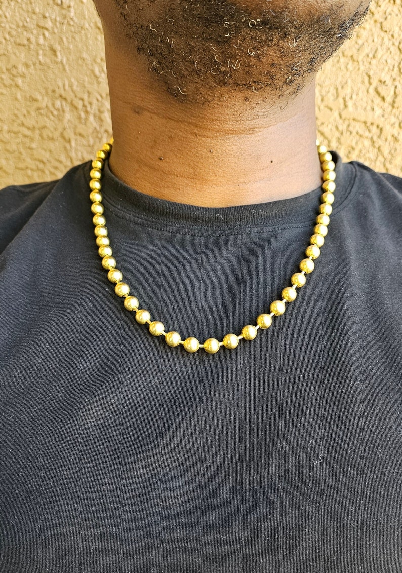 Op de afbeelding: Een gouden ketting met ronde kralen. De ketting wordt gedragen door een persoon met een zwart shirt.
