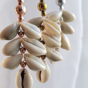 Peut inclure: Trois paires de boucles d'oreilles pendantes avec un motif de coquillage. Chaque boucle d'oreille présente une couleur de métal différente : cuivre, or et argent. Les boucles d'oreilles sont ornées de petites perles et de plusieurs coquillages.