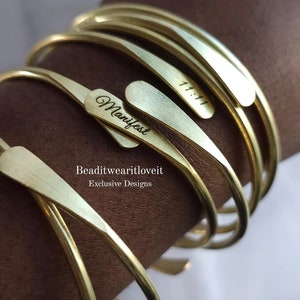 May include: A set of five gold-toned metal bangles with engraved text. The text on the bangles reads "Manifest" and "11:11". The bangles are stacked on top of each other and are worn on a person's wrist.