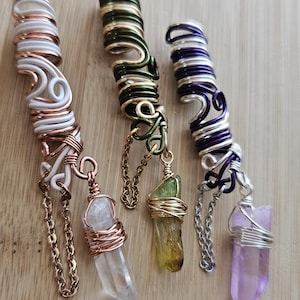 Op de afbeelding: Drie handgemaakte haaraccessoires met draadgewikkelde kristallen hangers. Eén is wit en koper, een andere is groen en goud, en de derde is paars en zilver. Elk heeft een ketting en een ander kristal.