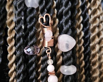 May include: Close-up of hair accessories, including a copper-colored spiral hair wrap with a heart design, and various pink gemstone beads. The hair is a mix of black and brown twists.