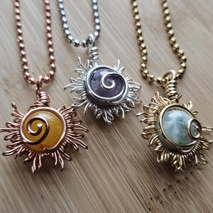 May include: Three sun-shaped pendant necklaces with different colored gemstones. The pendants are made of wire and are attached to ball chains. The gemstones are yellow, purple, and green. The necklaces are displayed on a wooden surface.