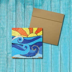 Puede incluir: Una tarjeta con un diseño de mosaico de un sol poniéndose sobre el océano. Las olas son azules y blancas, y el sol es amarillo, naranja y rojo. La tarjeta está sobre un fondo de madera azul.