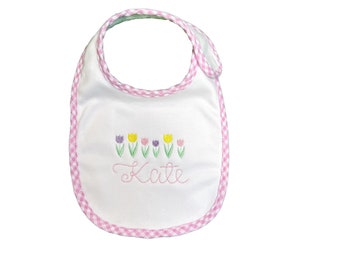 May include: White baby bib with a pink and white gingham trim. The bib has an embroidered design of a row of colorful tulips and the name "Katie".