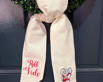 Op de afbeelding: Witte stoffen sjaal met rode borduurwerk. Het borduurwerk zegt "Roll Tide" en toont een mascotte van de Alabama Crimson Tide.