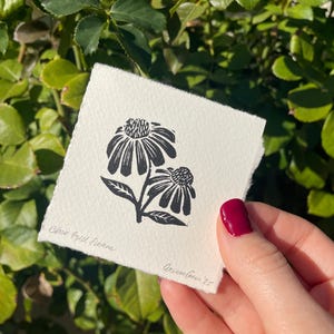May include: A black and white linocut print of two Black-Eyed Susan flowers on a square piece of paper. The print is held in a hand with red nail polish. The text "Black Eyed Susans" and "AriannaGreen '25" are visible.