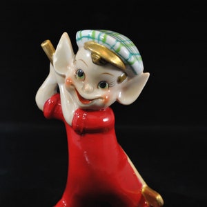 May include: A vintage ceramic figurine of a golf-playing elf. The elf is dressed in a bright red outfit with a white collar and a plaid hat. The figurine has a cheerful expression and is holding a golf club.