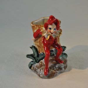 May include: A ceramic figurine of an elf holding a log. The elf is wearing a red hat and has a white beard. The log is brown and has green leaves on it.