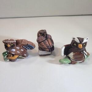 May include: Three ceramic owl figurines in various poses. The owls are primarily brown, with accents of white, blue, and green. Each owl is perched on a small, green base. The figurines have detailed feather patterns and expressive eyes.