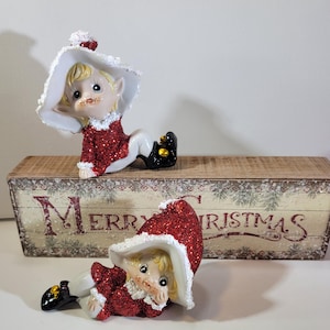 May include: Two Christmas elf figurines. Each elf is wearing a red glittery top, white pants, and black shoes with gold accents. They have blonde hair and are wearing white hats. The figurines are on a wooden box with "MERRY CHRISTMAS" printed on it.