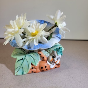 May include: A ceramic planter shaped like a cartoon character with a blue and white hat, holding white daisies. The planter is decorated with green leaves and orange spots.