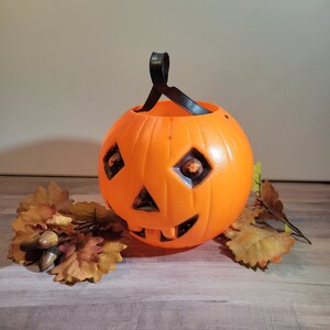 May include: Orange plastic pumpkin shaped Halloween trick-or-treat bucket with a black handle. The bucket has a carved jack-o'-lantern face with a toothy grin. The bucket is sitting on a wood surface with brown leaves and acorns.