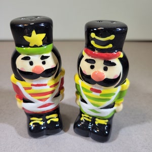 May include: A pair of ceramic salt and pepper shakers shaped like toy soldiers. The soldier on the left is wearing a red, yellow and white uniform with a green hat and a yellow star. The soldier on the right is wearing a green, yellow and white uniform with a red hat.