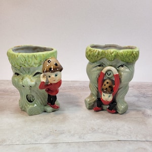 May include: Two ceramic planter pots shaped like tree stumps with green and brown paint. Each pot features a small elf figurine with a red shirt and black trousers. One elf is holding a mushroom and the other is peeking around the side of the pot.