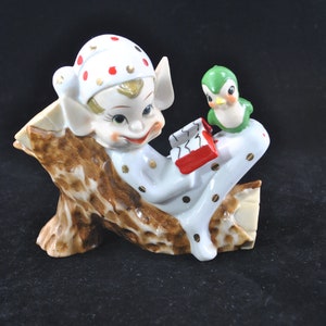 May include: A white ceramic figurine of an elf sitting on a tree stump with a red book and a green bird perched on the stump. The elf is wearing a white and red hat with gold trim and a white and gold outfit with red and gold polka dots.