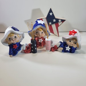Puede incluir: Tres figuras de elfos patrióticos con trajes y sombreros rojos, blancos y azules. Cada elfo tiene una estrella en el pecho y sostiene un pequeño fuegos artificiales. Una estrella de madera con un diseño de bandera está en el fondo.