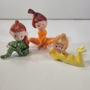 Puede incluir: Tres figuras de elfos de cerámica en varias poses. Uno está sentado en verde, otro en naranja y el tercero está acostado en amarillo. Cada elfo tiene el pelo rojo y una expresión sonriente.