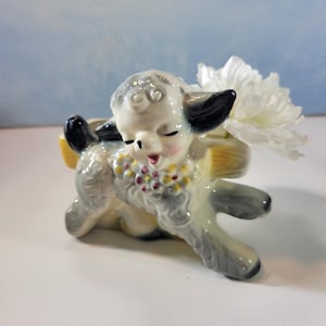 May include: A ceramic lamb figurine with a yellow basket on its back, holding a white flower. The lamb is painted in shades of gray, black, and pink, with a decorative floral collar. The figurine is on a white surface against a blue background.