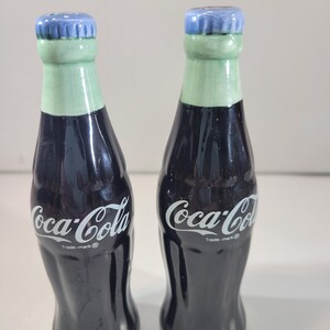K&ouml;nnte beinhalten: Ein Paar Salz- und Pfefferstreuer aus Keramik in Form von Coca-Cola-Flaschen. Die Flaschen sind dunkel mit wei&szlig;em "Coca-Cola"-Schriftzug, gr&uuml;nen H&auml;lsen und blauen Flaschendeckeln. Der Boden der Flaschen ist ebenfalls gr&uuml;n.