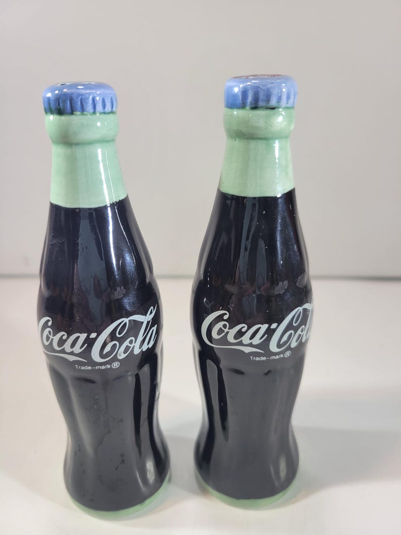 K&ouml;nnte beinhalten: Ein Paar Salz- und Pfefferstreuer aus Keramik in Form von Coca-Cola-Flaschen. Die Flaschen sind dunkel mit wei&szlig;em "Coca-Cola"-Schriftzug, gr&uuml;nen H&auml;lsen und blauen Flaschendeckeln. Der Boden der Flaschen ist ebenfalls gr&uuml;n.