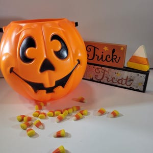 May include: An orange plastic jack-o'-lantern bucket with a smiling face, black eyes, and a black mouth. Candy corn is scattered around the bucket. A wooden sign says "Trick or Treat". A wooden candy corn decoration is also present.