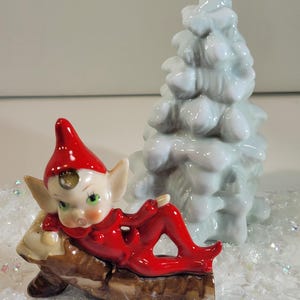 Puede incluir: Una figura de elfo navideño de cerámica con un atuendo y sombrero rojos, reclinada sobre un tronco. Un árbol de Navidad de cerámica azul claro está detrás del elfo. La escena está sobre una superficie blanca con purpurina.